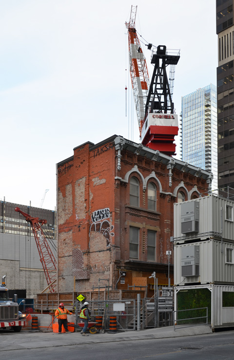 old brick three storey building stands alone by a construction site as new development goes on around it. large crane in the background 