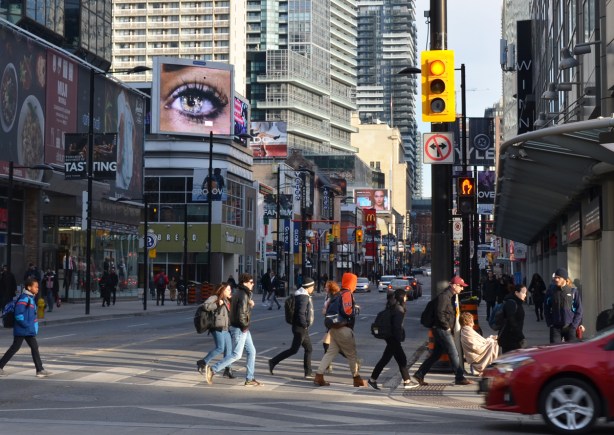 people cross Yonge street on the north side of Dundas, a big picture of an eye is on a billboard looking down over the street 