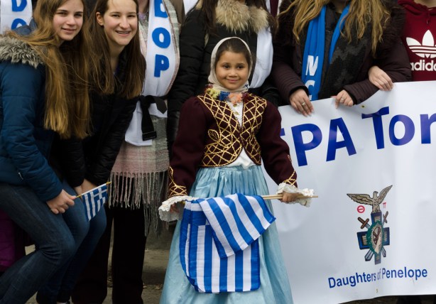 3 girls posing with a banner, the younger girl holds a Greek flag, they are with a banner representing the Daughters of Penelope