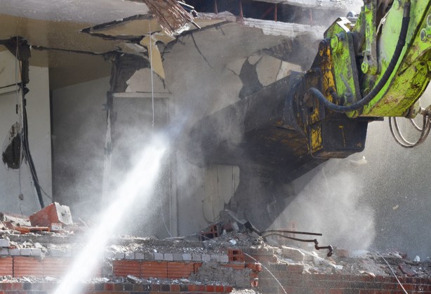 close up of dust and debris as workmen spray water as a machine arm pulls apart a building that is in the process of being demolished