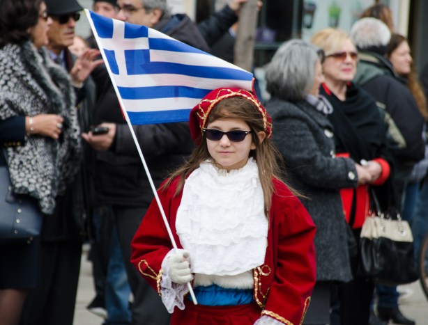 a girl in red jacket and cap, dark sunglasses, and holding a Greek flag, walks in a parade, with people on the sidewalk watching in the background. 