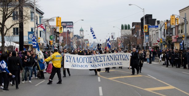 a police man in yellow jacket directs a group of marchers in the Greek Independence Day parade on the Danforth. The group is walking with a large banner with words written in Greek 