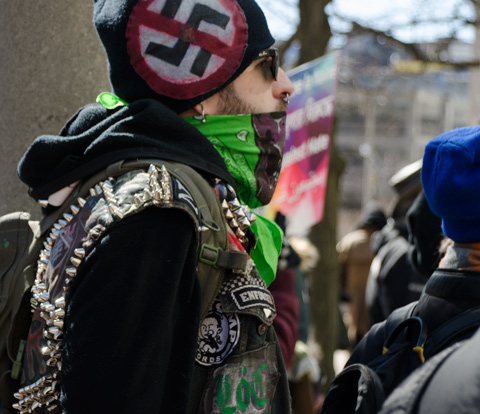 a man wears a green and brown bandana over the bottom part of his face and a black hat with an anti-nazi symbol on it. 