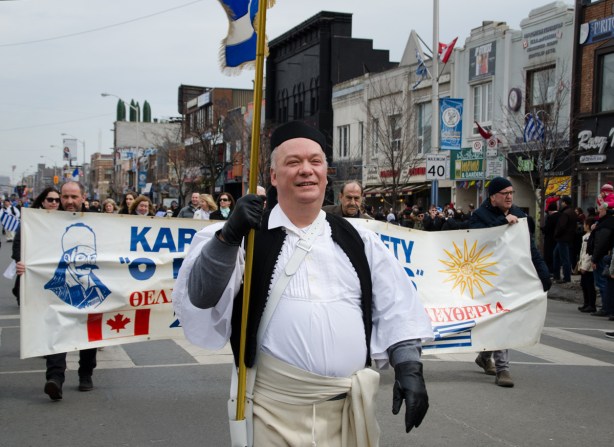 a man in traditional Greek clothing carries a flag in a parade, he is in front of a group carrying a banner