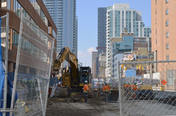 cain link fence and gate is open, construction crew in the middle of Eglinton Ave (at Mt Pleasant) is working with a digger, hole in the ground