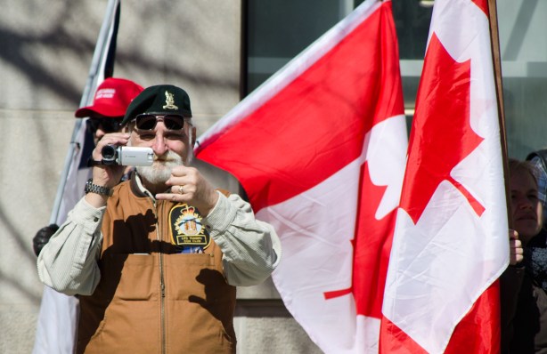 man filming crowd at a protest, two Canadian flags beside him 