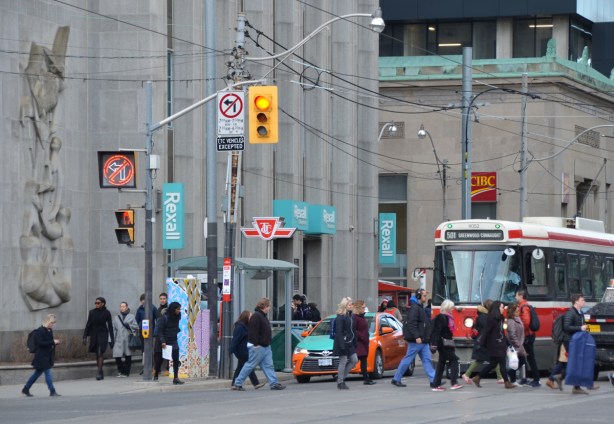 people crossing the street in front of a streetcar that is waiting for a red light. At thewest side of intersection of Queen and University