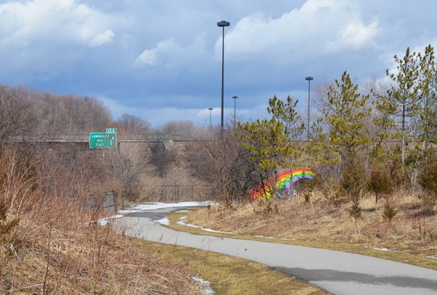 East Don Trail winds towards the bridge that is painted with a rainbow, some traces of the Don Valley Parkway like a green exit sign for Lawrence Avenue, and a couple of tall light stands. 