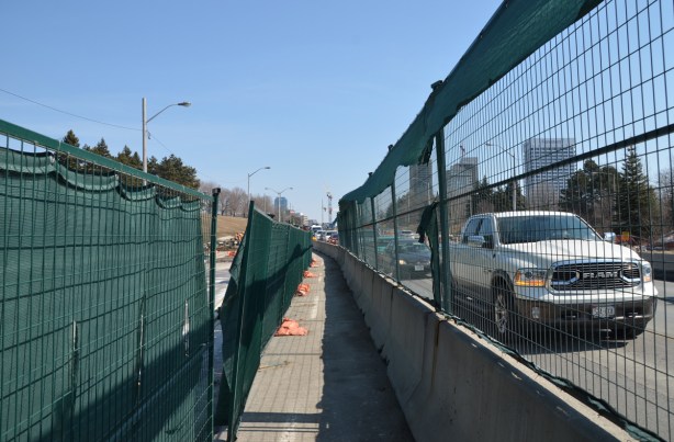 green netting and fencing on both sides of a narrow sidewalk running between construction and traffic. 