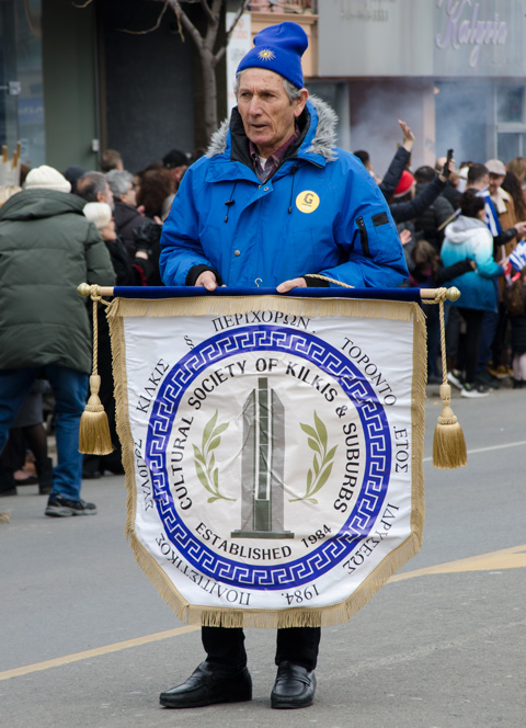 a man in a blue parka carries a banner in a parade, a small flag-like banner for the Cultural Society of Kilkis 