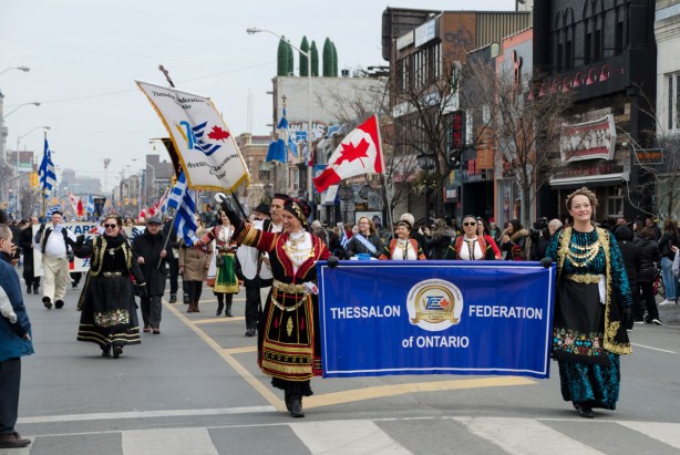 women in traditional Greek costume carry a banner of the Thessalon Federation of Ontario, a blue banner, in a parade, while others walk behind the banner