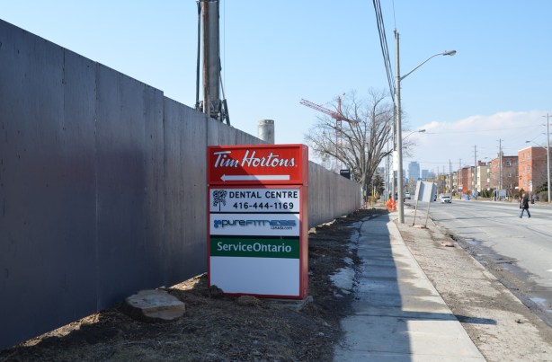 red and white tim hortons sign with an arrow pointing left at a long grey fence around a construction site, sidewalk, street, and low rise buildings on the right, Eglinton Ave