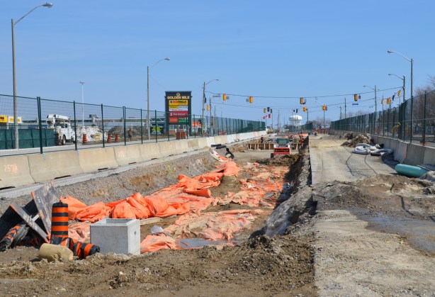 shallow but wide hole in the ground where new LRT tracks are being laid. construction in prep for the tracks, green fencing separates construction from traffic on both sides, 