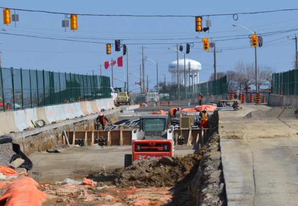 shallow but wide hole in the ground where new LRT tracks are being laid. construction in prep for the tracks, green fencing separates construction from traffic on both sides, water tower in the distance