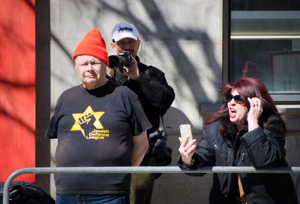 a woman with dyed auburn hair is making a video of herself at a protest on her cell phone, two men are also in the photo, one has an orange toque and a Jewish defence league T-shirt and the other has a white baseball cap that says Jesus is my boss as he takes pictures of the crowd on the other side of the protest 