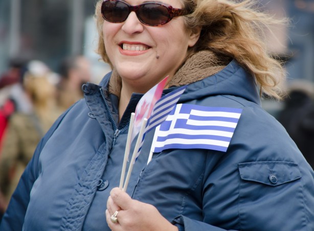 a woman with dark sunglasses and blue coat holds three small flags, two Greek and one Canadian 