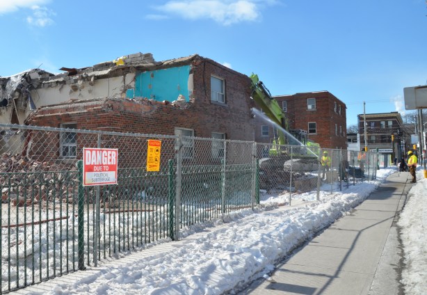 workmen spray water as a machine arm pulls apart a building that is in the process of being demolished - sidewalk view