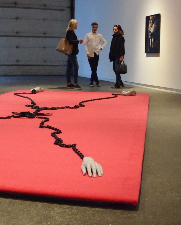 three people stand in a large open gallery space, looking at a large red piece on the floor, two hands, and two feet, fill the four corners, and a black harness connects them through the center, a photo is on the wall beside them - it is of entrails. It is the work of Chris Curreri