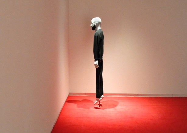 a mannequin top, of a bald man, on a frame with black loose fitting clothes, stands on a red carpets facing the corner, nothing else is in the room 