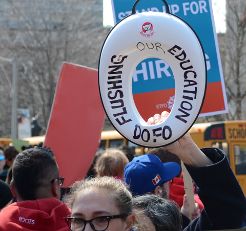 person holding a toilet seat protesting Doug Fords proposed cuts to education funding, words written on seat say DoFo flushing education