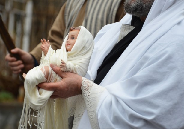a bearded priest carrying a baby doll wrapped in white swaddling clothes in a passion procession 