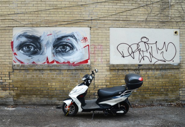 black and white realistic painting of a pair of eyes, larger than life, on a yellowish brick wall.  A small motorcycle is parked in front of the wall. 