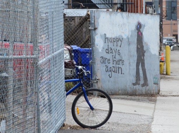 a blue bike leans against a chainlink fence, behind it is a piece of street art that says happy days are here again, with painting of a man walking 
