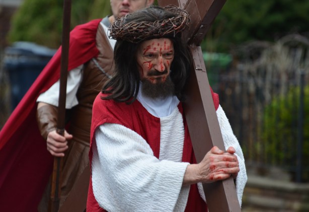 a man with a crown of thorns and blood on his face and carrying a large cross walks in a parade, with a man behind him dressed as a roman soldier 