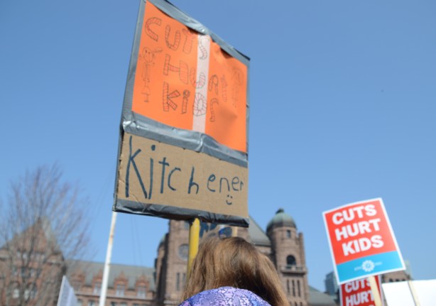 person holding a placard protesting Doug Fords proposed cuts to education funding, child on adult shoulders.