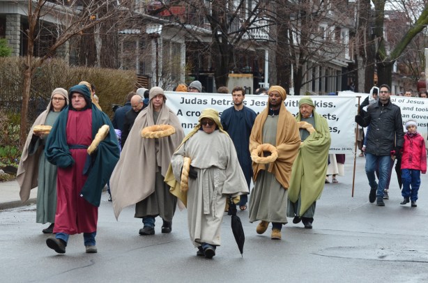 small group of men in a parade, wearing long robes and carrying bread in the shape of a large wreath, a banner is behind them that describes the betrayal of Jesus by Judas