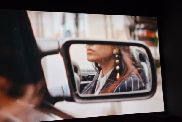 picture from a video, a woman's reflection in the side mirror of a car, long hair, long dangly ear rings 
