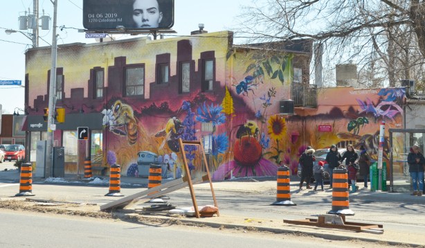 large mural on the side of a building, bus shelter and stop to the side with people waiting. Mural is by Nick Sweetman and features many bees and flowers, very large bees