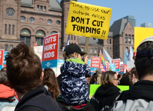 person holding a placard protesting Doug Fords proposed cuts to education funding, child on adult shoulders.Crowds at a protest at Queens Park, 