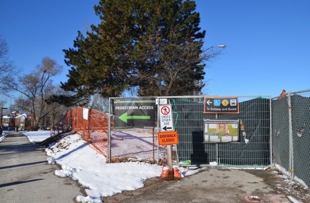 a sidewalk ends at a fence arond a construction site and pedestrians are diverted through a park to the left, signs on the fence directing traffic 