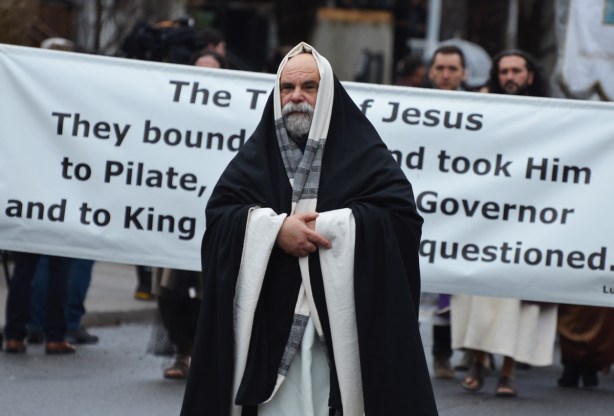 a man dressed as a priest in long black and white robes walks in a passion of christ procession in front of a large banner that has a bible verse from Luke 23 