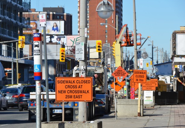 sidewalk, many orange construction signs cluttering the sidewalk, bus stop, traffic on the street, 