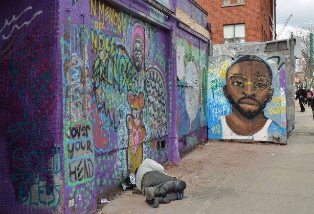 a man sleeping on the sidewalk in front of a wall covered with street art, graffiti and art in memory of Andre Alexander who was an artist who worked in Kensington
