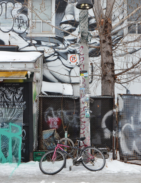 a bike locked to a tree on a sidewalk in front of a mural on a building, snow, 