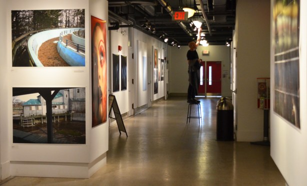 part of the third floor hallway gallery space at Artscape Youngplace with large colour photos tacked to the wall, a man on a ladder adjusts the lighting