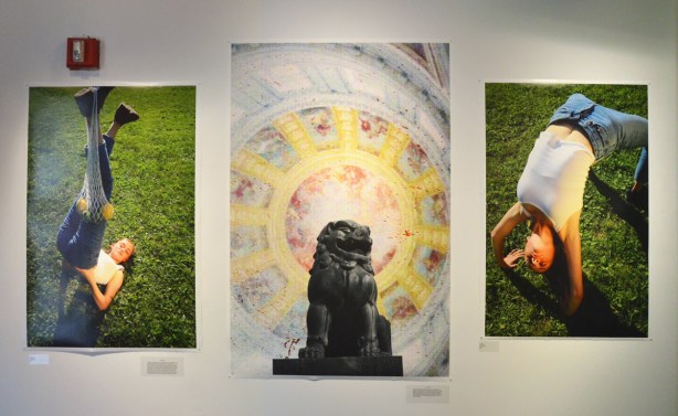 three photographs hanging on a wall. On either side is a picture of a woman in a pose, on green grass outside by Julia Kerrigan. In the middle is a black statue, Asian lion, in front of a light pastel coloured circular pattern