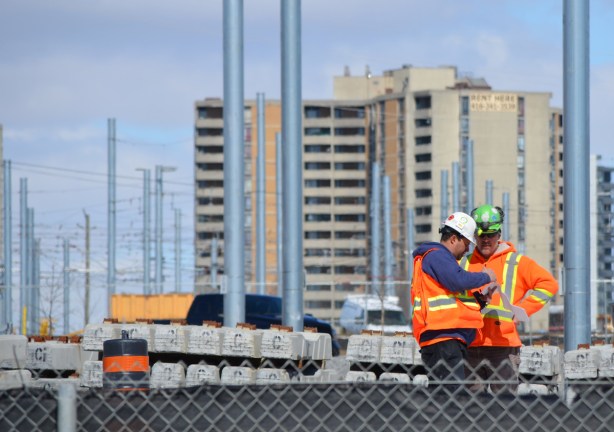 two workmen in hard hats and orange work vest look at paperwork on a construction site. 