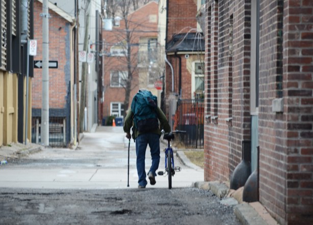 a man with a cane in one hand, and pushing a bike with other walks down an alley. he also has a backpack on his back