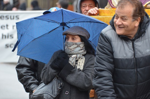 a woman bundled up in grey tam and scarf over the bottom of her face and carrying a blue umbrella, walking with some men in a passion of christ good friday procession 