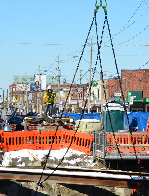 a workman in hard hat and yellow jacket stands on a pile of steel rods on the back of a flat bed truck with a crane lowering a steel beam into the ground in front of him