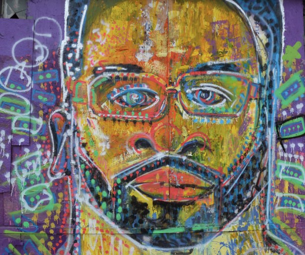 large painting of a face, street art, yellowish skin, small moustache and beard, glasses, eyes looking straight ahead