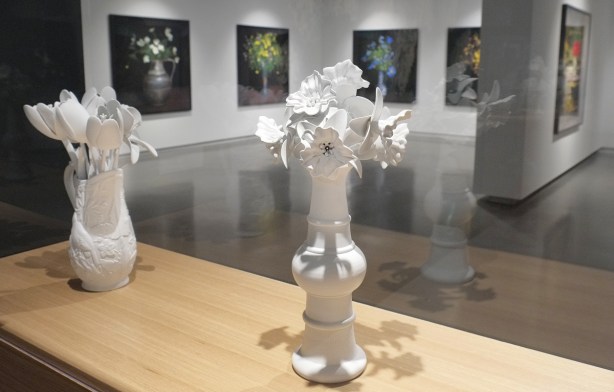 2 white 3 D printed sculptures of flowers in a vase, in a gallery, with large pictures, in colour, of bouquets of flowers in vases on tables