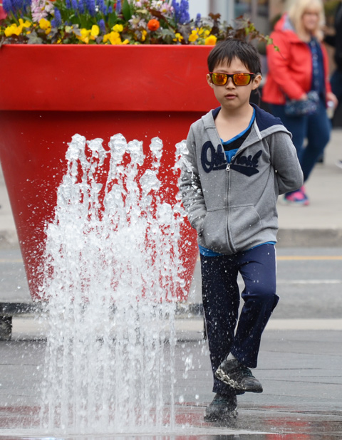 a young boy in sunglasses stands on one foot in front of a water fountain at Dundas square