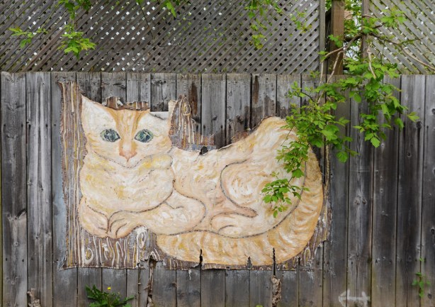 large painting of a yellow and white cat on canvas stapled to a wood fence, outdoors. 