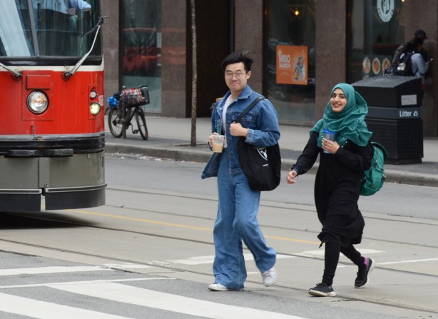 a couple crosses a street by a streetcar, an Asian man and a woman in a teal head scarf 