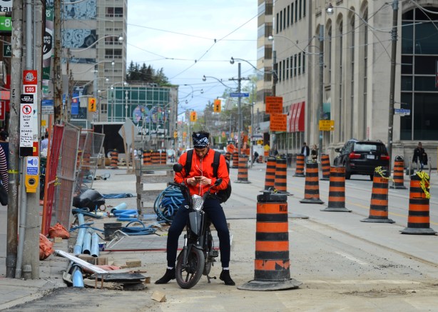 a cyclist with an otange shirt sits on his bike while looking at his phone. He's stopped behind an orange and black striped construction cone on the side of a street 